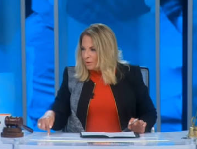 A blonde woman in an orange top and dark blazer sits at a news desk with papers and a gavel, set against a blue studio background.