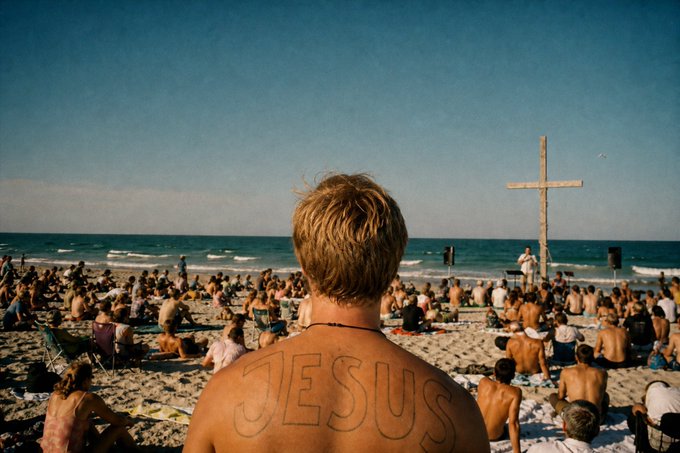 A photo from behind a blond-haired man with 'JESUS' written on his back, facing a crowded beach where a large wooden cross stands near the ocean.