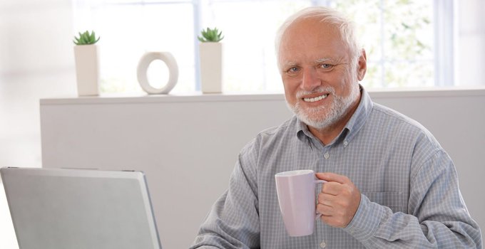 A smiling older man with a white beard holding a lavender mug while sitting in front of a laptop, with potted plants and a decorative ring on a shelf in the background.