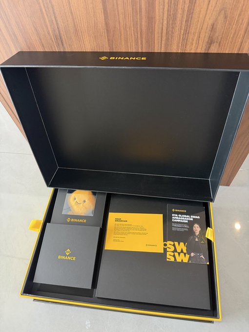 An open Binance premium gift box with yellow interior containing branded materials, a staff mountain photo card, and an 'FY25 Global Brand Ambassador Campaign' document.