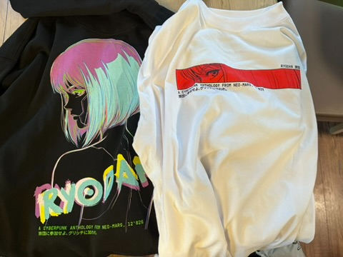 Two graphic t-shirts are displayed on a surface; the black one features an anime-style illustration with the text 'RYOAS' and 'CYBERPHUNK ANTOLOGY FOR NEO-MARS', while the white one shows a red graphic with an eye design.