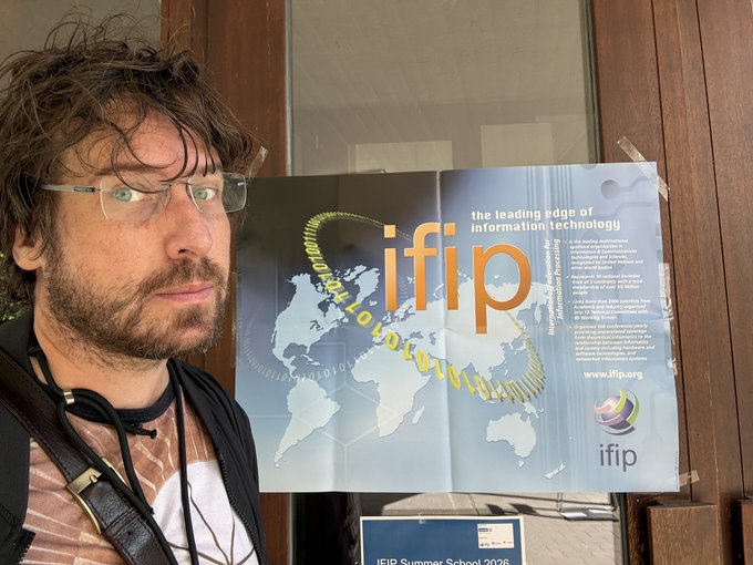 A bearded man with glasses poses next to an IFIP (International Federation for Information Processing) promotional poster advertising 'the leading edge of information technology' for their Summer School 2026.