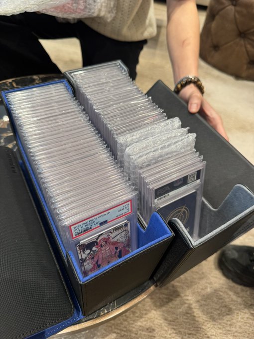 A person holding an open box filled with neatly stacked rows of graded trading cards in protective slabs, including a Pokémon card featuring a pink character with a PSA grade label.