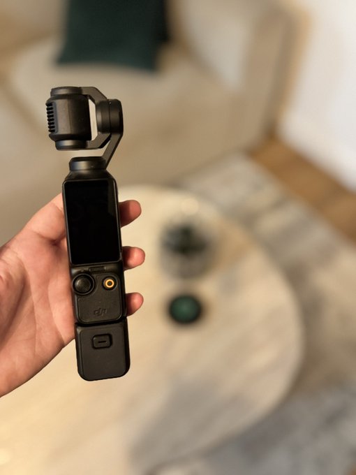 A hand holding a DJI Osmo Pocket 3 handheld gimbal camera with its small screen visible, photographed in a living room with a coffee table in the blurred background.