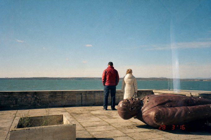 A photograph dated '28 3 '16' showing two people standing by a stone wall overlooking a body of water, with a large rusted metal statue lying on the ground nearby and a water fountain visible in the distance.