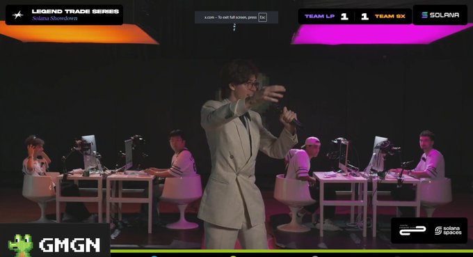 A broadcast screenshot of the 'LEGEND TRADE SERIES' esports event showing a host in a suit holding a microphone, with players at desks and a scoreboard showing 'TEAM LP 1 - 1 TEAM GX'.