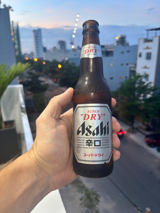 A hand holds up a bottle of Asahi Super "DRY" beer against a city skyline at dusk. The "never work again" caption is humorously contrasted with enjoying a casual evening drink, suggesting that successful crypto investing might fund a relaxed lifestyle rather than actual early retirement.