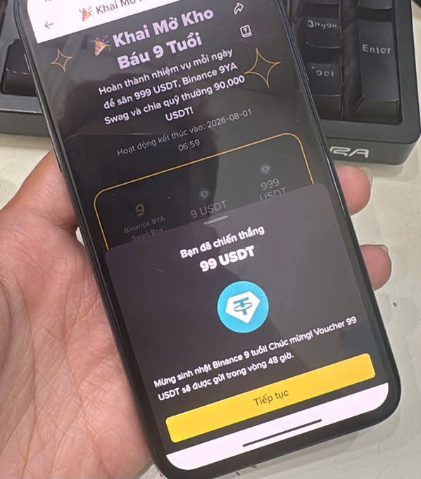 A hand holding a smartphone displaying a Binance app screen titled 'Khai Mở Kho Báu 9 Tuổi' in Vietnamese, celebrating Binance's 9th anniversary with a 99 USDT reward voucher.