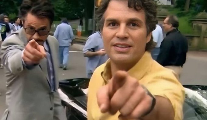 Two men point directly at the camera in an outdoor street setting, one in a yellow shirt and the other in a tan jacket with sunglasses.
