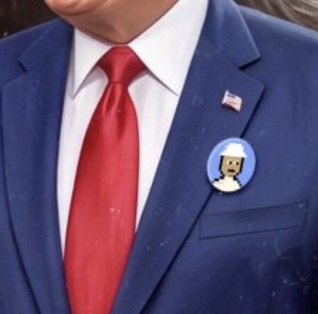A close-up shows a person in a blue suit with a red tie and an American flag pin, wearing a circular button featuring a shocked emoji wearing a hard hat.