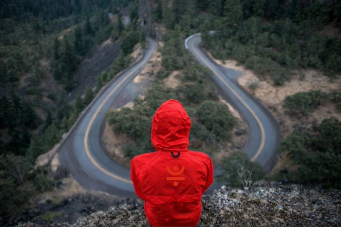 A person wearing a bright red hooded jacket with a small logo sits facing away from the camera, looking down at a winding paved road in a mountainous, forested landscape.