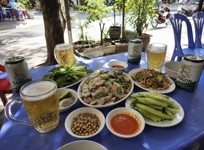 An outdoor table set for a Vietnamese meal features multiple dishes including meat, vegetables, cucumbers, peanuts, and chili sauce, accompanied by mugs of beer and cans of Bia Saigon.
