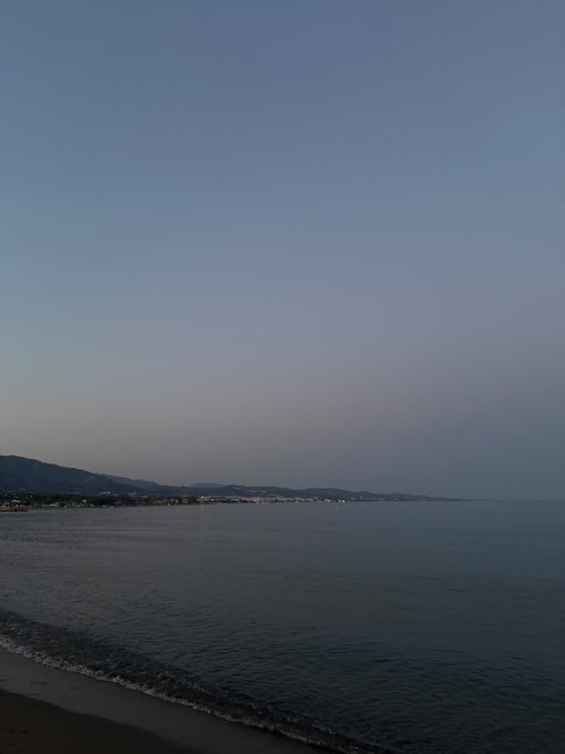 A serene view of the sea at dusk shows calm blue water, a distant coastline with mountains, and a clear gradient sky from pale to dark blue.