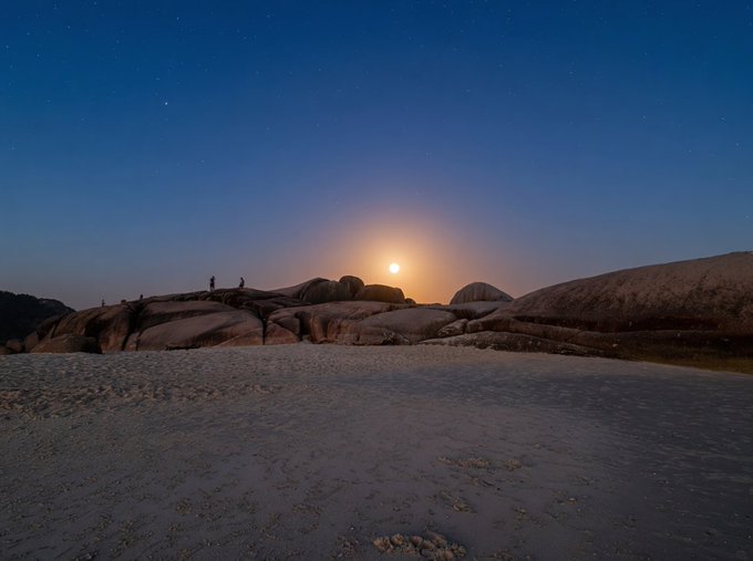 A nighttime landscape showing a glowing moon rising over rocky terrain, with two small silhouetted figures standing on the rocks and a sandy beach in the foreground.
