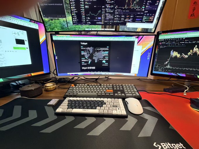A multi-monitor cryptocurrency trading setup displaying Bitget charts on several screens, featuring a mechanical keyboard, a white ergonomic split keyboard, a mouse, and a Bitget-branded desk mat.
