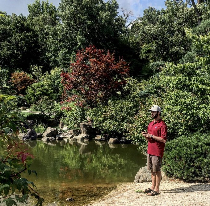 A man wearing a red shirt and cap stands at the edge of a pond in a lush garden, holding a phone, surrounded by trees and greenery.