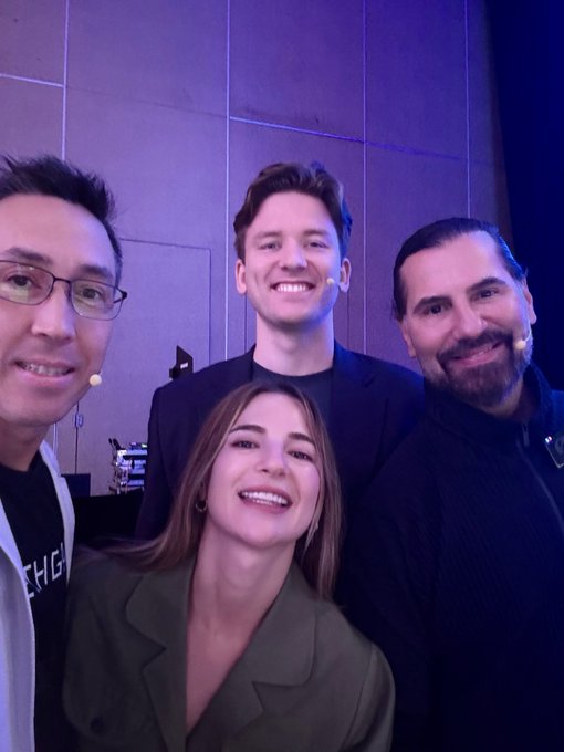A group selfie of four people smiling together at what appears to be a tech event, including an Asian man in glasses, a young man with a microphone headset in a suit, a laughing young woman, and a bearded man in a dark sweater.