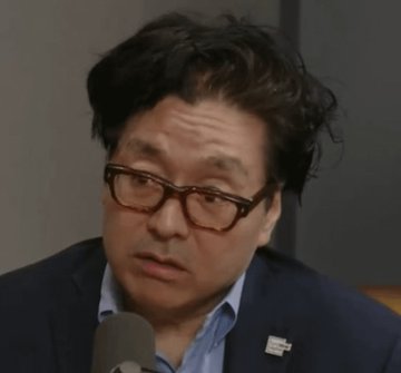 The image features a man wearing glasses and a suit jacket with his head tilted back as if he's speaking or listening intently. The background appears to be a gray wall, which contrasts with the man's attire. There are no other objects or people in the image besides the man and the gray wall.