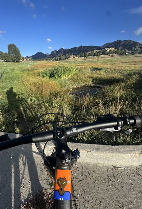 First-person view from a bicycle handlebar looking out over a grassy field with mountains in the distance under a clear blue sky.