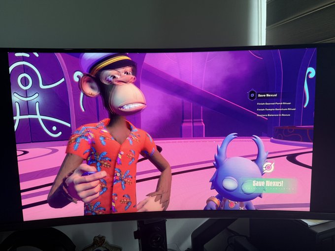 The image shows a computer monitor displaying an animated video game scene featuring two characters, one wearing a red shirt with a hat and the other dressed as a purple alien. The character in the red shirt appears to be giving advice or instructions to the purple alien character on the screen.
