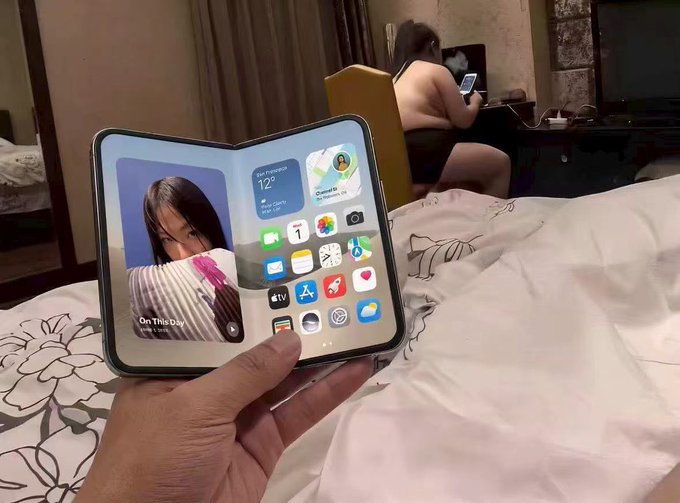 A first-person view of a hand holding a foldable smartphone (appearing to be a Pixel Fold) displaying the home screen with widgets and apps, while another person sits on a bed in the background using their phone.