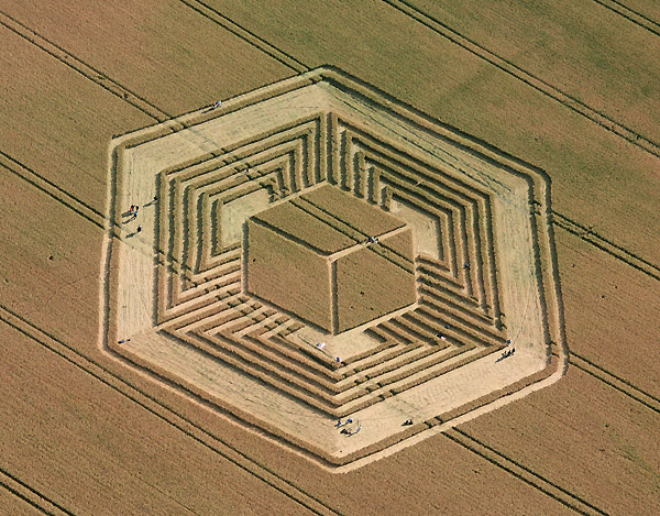 Aerial view of a large hexagonal earthwork with a central cube and concentric rings carved into a grassy field.