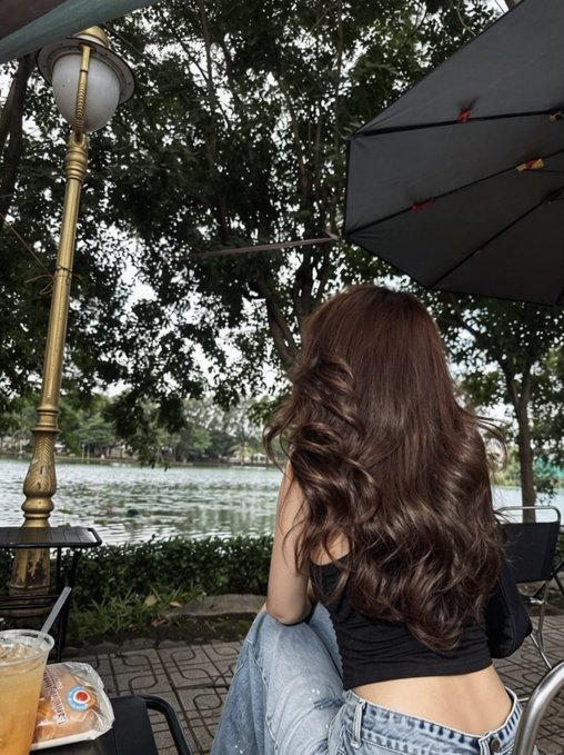 A person with long, wavy brown hair sits from behind at an outdoor cafe, wearing a black top, with a lake, trees, a lamp post, and an umbrella visible in the scene.