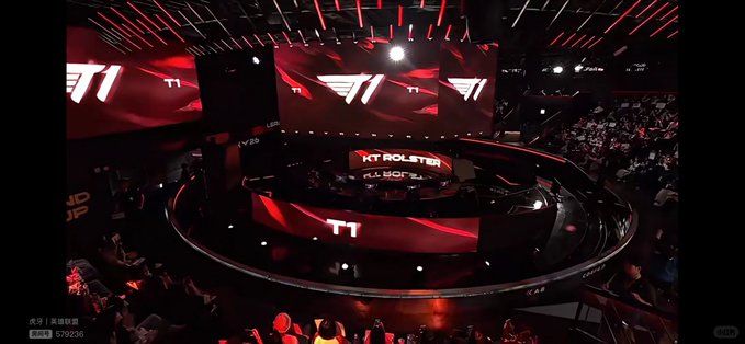 An esports arena stage displays the T1 logo prominently on large screens, with a 'KT Rolster' advertisement visible and a crowd of spectators in the foreground.