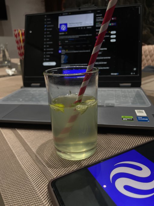 A glass of yellow drink with a red and white striped straw sits in front of an open laptop displaying a website, with a phone showing a blue app logo in the foreground.