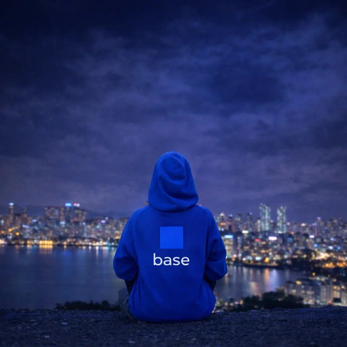 A person wearing a blue hoodie with the "base" logo sits on a ledge overlooking a city skyline at night.