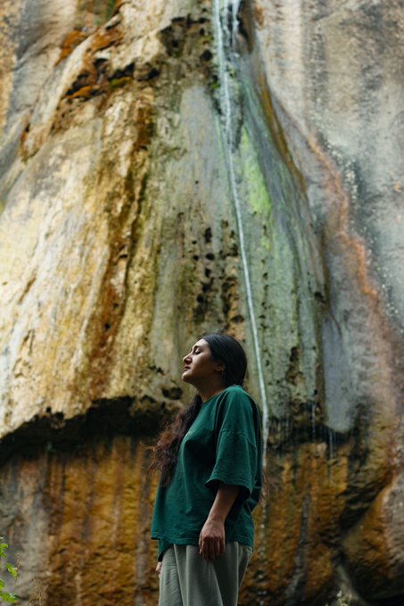 A woman with long dark hair stands in profile wearing a green t-shirt, looking up at a thin waterfall trickling down a textured rock face.