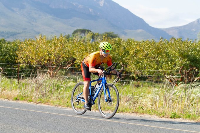 A cyclist in a yellow and red jersey and green helmet riding a blue Giant road bike on a paved road, with green vineyards and mountains in the background.