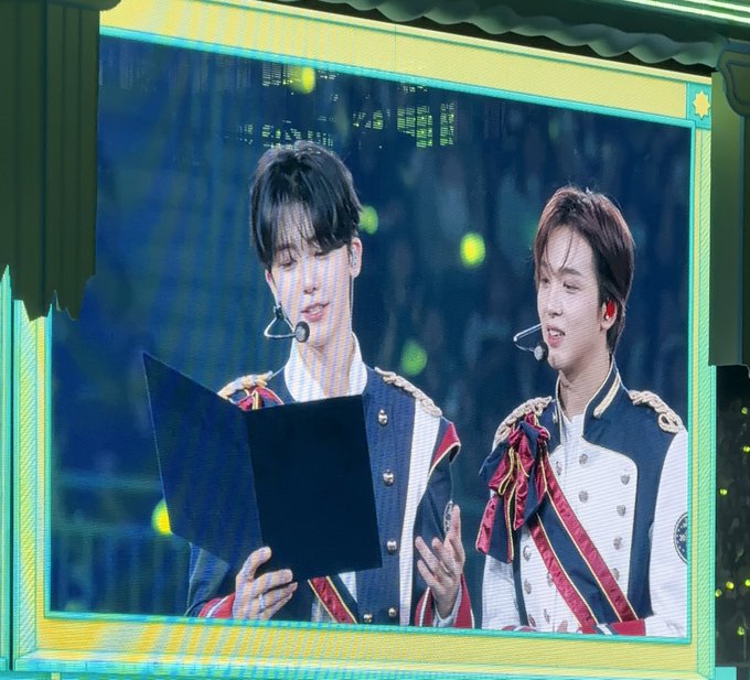 Two performers in ornate navy and red military-style jackets with epaulettes and headset microphones are shown on stage, with one reading from a black folder while both appear to sing or speak. The image appears to be displayed on a large screen during a concert or live performance event.