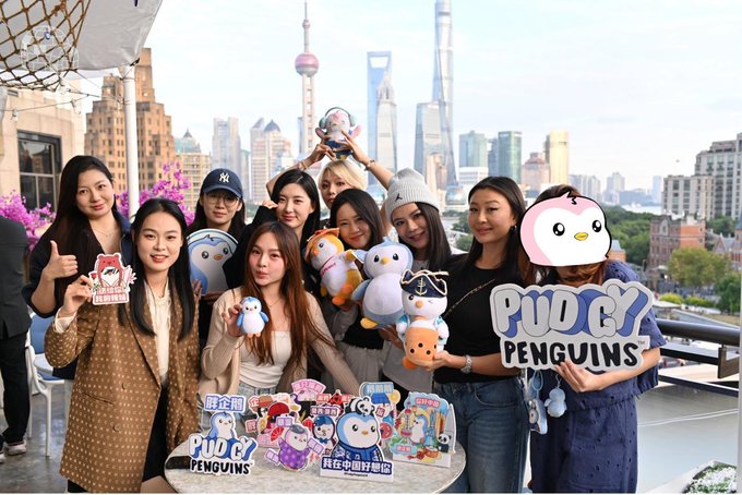 A group of women posing with Pudgy Penguins merchandise and plush toys on a rooftop overlooking the Shanghai skyline.