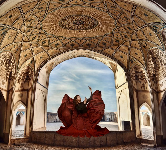 A woman in a flowing red dress poses dramatically inside an ornate Persian architectural archway with an intricate patterned ceiling.