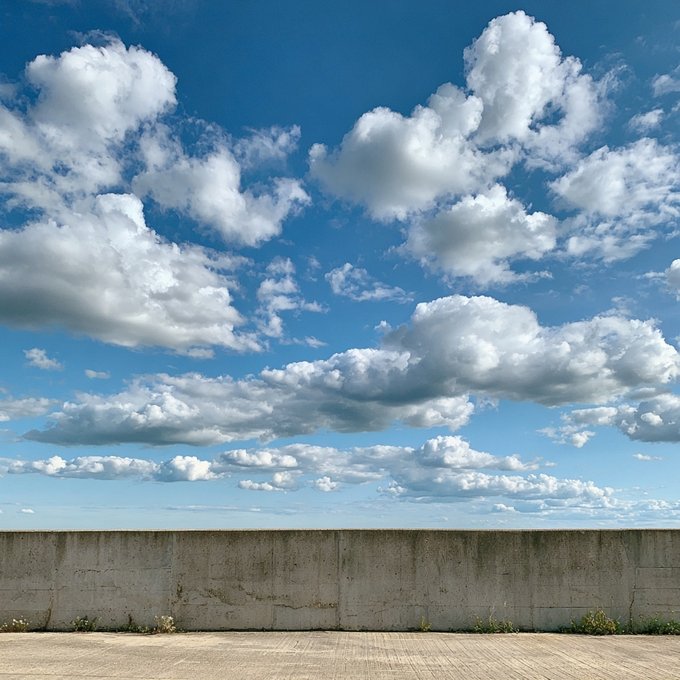 A scenic view of a bright blue sky filled with puffy white clouds, seen above a low concrete wall and a paved surface.