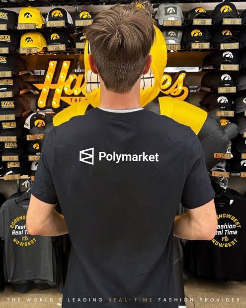 A person with short brown hair wearing a black 'Polymarket' t-shirt stands with their back to the camera in a store displaying various hats and apparel, with 'THE WORLD'S LEADING REAL-TIME FASHION PROVIDER' text at the bottom.