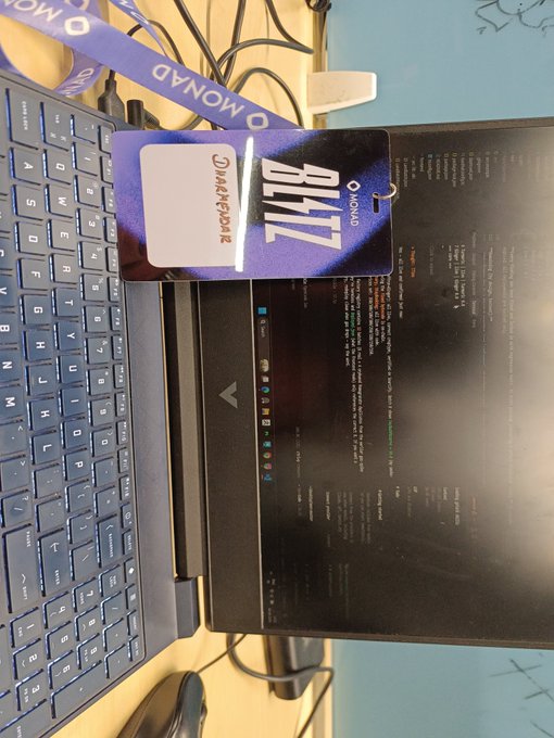 An overhead view of a desk showing a laptop keyboard, an LG monitor displaying code or terminal text, and a purple 'OMONAD BLITZ' conference badge.