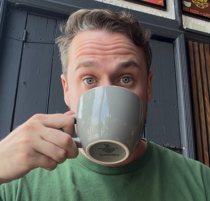 A man in a green shirt drinks from a grey 'Threshold' mug, looking over the rim at the camera.