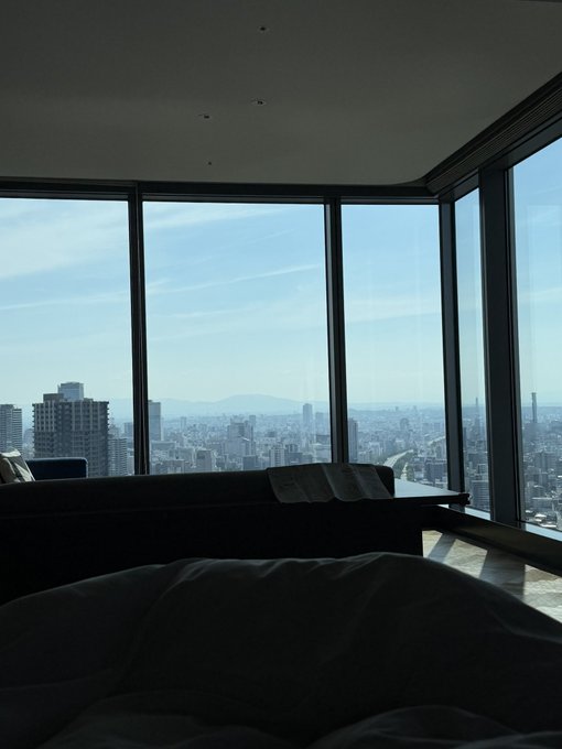 A view from a high-rise hotel room window looking out over a city skyline and distant mountains under a clear blue sky, with a bed visible in the foreground.