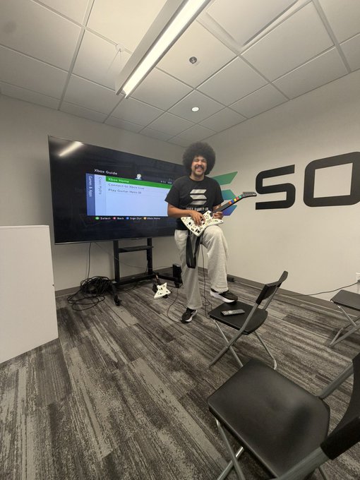 A person with an afro playing a white electric guitar while jumping on a chair in what appears to be a gaming venue with 'SQ' on the wall.