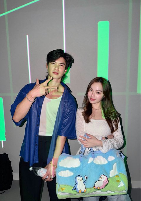 A young man in a blue button-up over a white tee poses with a peace sign next to a young woman in a white top holding a fuzzy blue penguin-themed bag at what appears to be a concert or event.