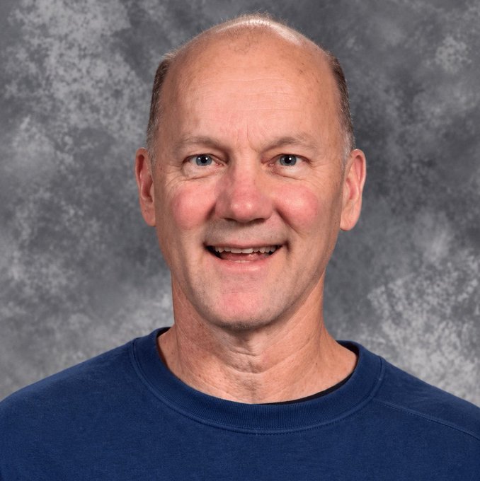 A professional headshot photograph of a smiling, balding middle-aged man with light eyes wearing a dark blue t-shirt against a mottled grey studio backdrop.
