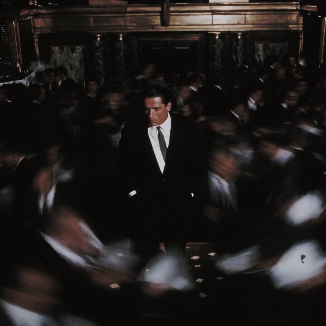 The image shows a sharply-dressed man in a black suit and white tie standing still amid a swirling, motion-blurred crowd in a dark wood-paneled room, evoking a cinematic, noir atmosphere. The accompanying post's cryptic "Big reveal soon. Notifs on" suggests this enigmatic figure may be a teaser image for an upcoming announcement or project.