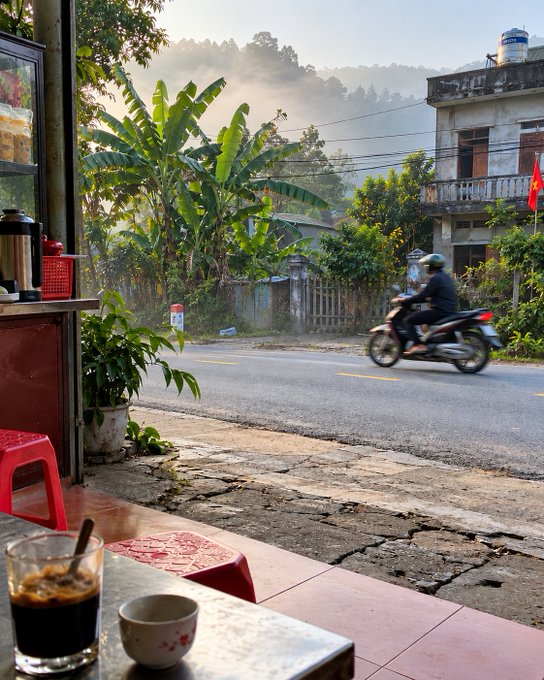 A view from a street-side cafe looking out onto a road where a person rides a motorcycle, with misty mountains and a building flying a red flag in the background.