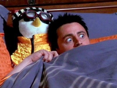 A still image from the movie 'Elf' showing Buddy the Elf (Will Ferrell) in bed with wide-eyed excitement next to a penguin wearing pajamas and goggles.