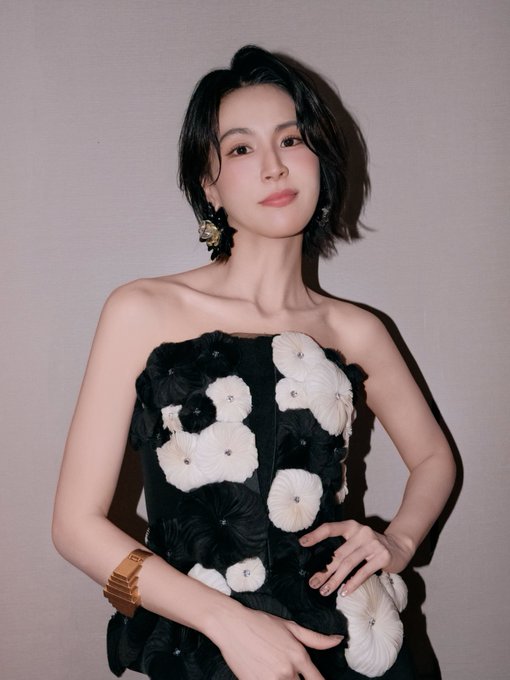 A portrait of a woman with short black hair wearing a black strapless dress adorned with 3D white and black floral appliqués, accessorized with gold floral earrings and a chunky gold cuff bracelet.
