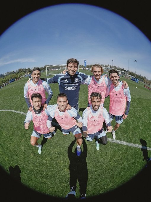 A fisheye-lens team photo of seven soccer players on a green field, with the back player wearing a navy Adidas jersey featuring the Coca-Cola logo and the rest in pink training bibs.