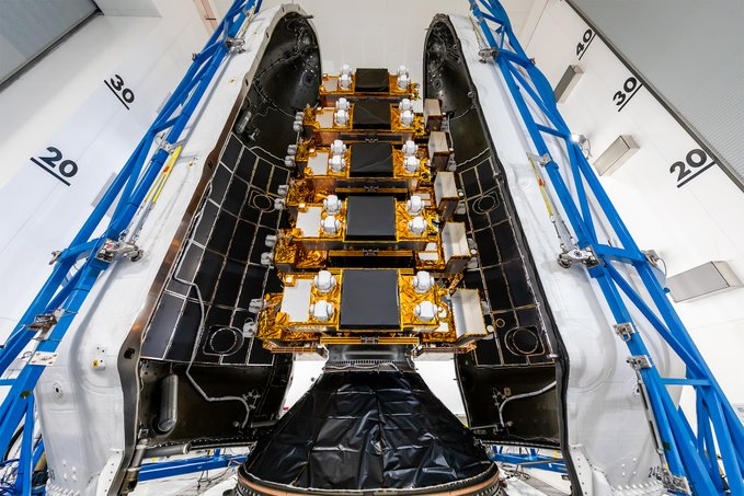 A top-down view of a spacecraft payload bay revealing gold-foiled satellites stacked on a central platform, surrounded by blue support structures inside a clean room facility.