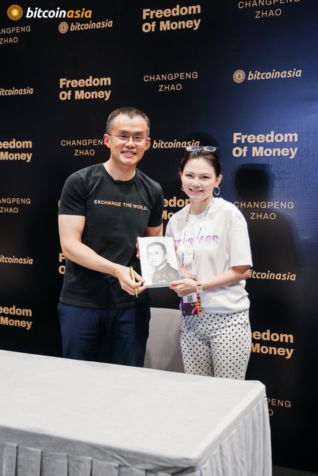 A man in a black t-shirt and a woman in a white t-shirt and polka dot pants pose together at a "bitcoin asia" / "CHANGPENG ZHAO" / "Freedom Of Money" event, holding up a portrait sketch.
